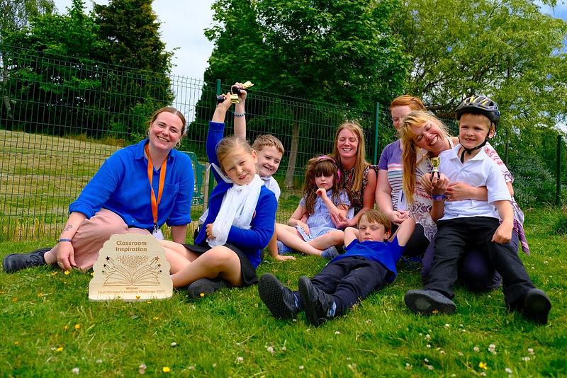 A group of pupils and their teachers celebrating receiving their First Minister's Reading Challenge trophy. They are sitting together on the grass in the playground.